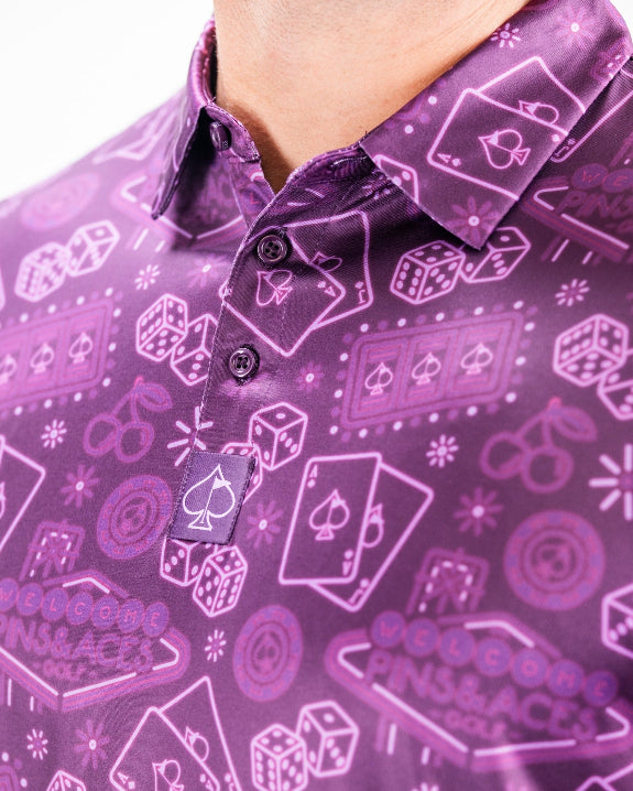Men’s purple casino golf polo by Pins & Aces featuring an all-over pattern of playing cards, dice, poker chips, slot machines, and Las Vegas-inspired graphics.