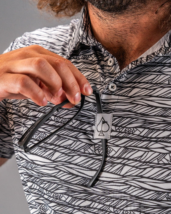 Men’s Fragment Black golf polo by Pins & Aces featuring an all-over black, white, and gray abstract line pattern with short sleeves and an athletic performance fit.