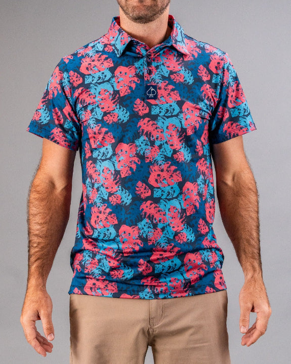 Men’s Painted Monstera Blue golf polo by Pins & Aces featuring coral and aqua tropical monstera leaves on a deep blue background with short sleeves and an athletic performance fit.