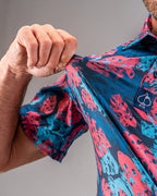 Men’s Painted Monstera Blue golf polo by Pins & Aces featuring coral and aqua tropical monstera leaves on a deep blue background with short sleeves and an athletic performance fit.