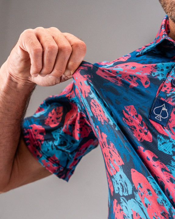 Men’s Painted Monstera Blue golf polo by Pins & Aces featuring coral and aqua tropical monstera leaves on a deep blue background with short sleeves and an athletic performance fit.