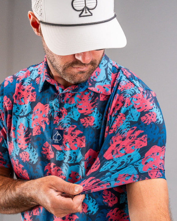 Men’s Painted Monstera Blue golf polo by Pins & Aces featuring coral and aqua tropical monstera leaves on a deep blue background with short sleeves and an athletic performance fit.