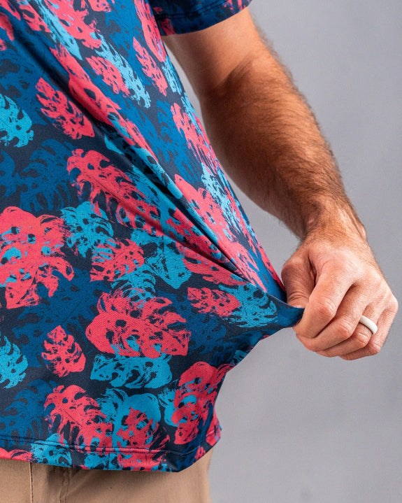 Men’s Painted Monstera Blue golf polo by Pins & Aces featuring coral and aqua tropical monstera leaves on a deep blue background with short sleeves and an athletic performance fit.