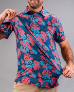 Men’s Painted Monstera Blue golf polo by Pins & Aces featuring coral and aqua tropical monstera leaves on a deep blue background with short sleeves and an athletic performance fit.