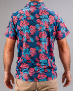 Men’s Painted Monstera Blue golf polo by Pins & Aces featuring coral and aqua tropical monstera leaves on a deep blue background with short sleeves and an athletic performance fit.