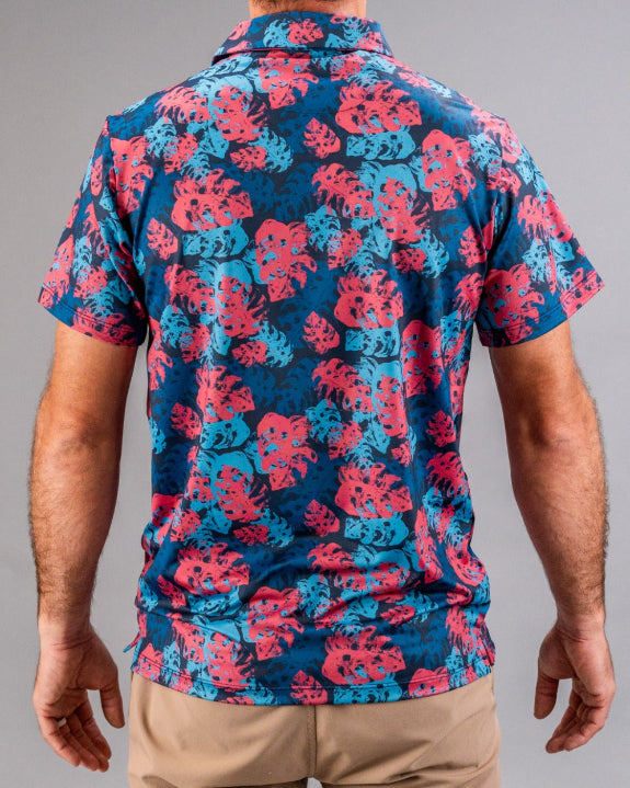 Men’s Painted Monstera Blue golf polo by Pins & Aces featuring coral and aqua tropical monstera leaves on a deep blue background with short sleeves and an athletic performance fit.
