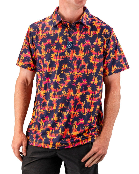 Men’s Palm Drip Dusk golf polo by Pins & Aces featuring navy palm tree silhouettes over a vibrant orange, pink, and purple tropical sunset print with an athletic fit.