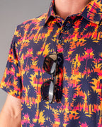 Men’s Palm Drip Dusk golf polo by Pins & Aces featuring navy palm tree silhouettes over a vibrant orange, pink, and purple tropical sunset print with an athletic fit.