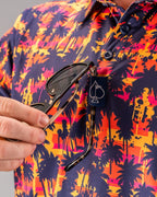 Men’s Palm Drip Dusk golf polo by Pins & Aces featuring navy palm tree silhouettes over a vibrant orange, pink, and purple tropical sunset print with an athletic fit.