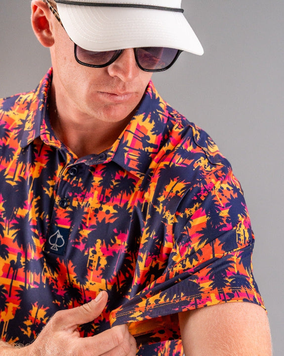 Men’s Palm Drip Dusk golf polo by Pins & Aces featuring navy palm tree silhouettes over a vibrant orange, pink, and purple tropical sunset print with an athletic fit.