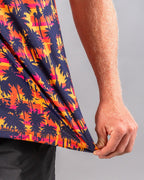 Men’s Palm Drip Dusk golf polo by Pins & Aces featuring navy palm tree silhouettes over a vibrant orange, pink, and purple tropical sunset print with an athletic fit.