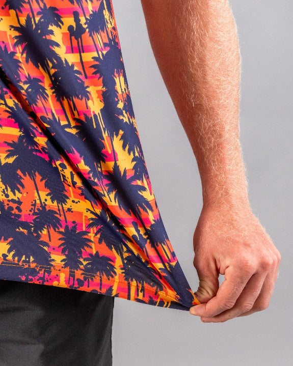 Men’s Palm Drip Dusk golf polo by Pins & Aces featuring navy palm tree silhouettes over a vibrant orange, pink, and purple tropical sunset print with an athletic fit.