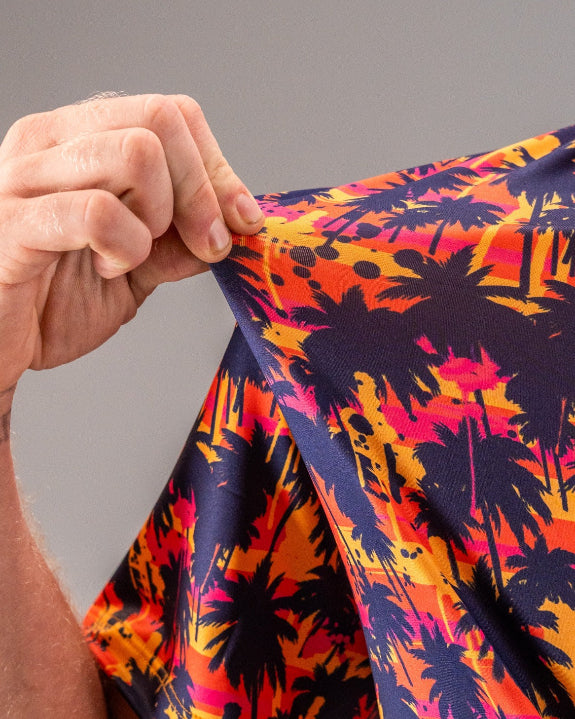 Men’s Palm Drip Dusk golf polo by Pins & Aces featuring navy palm tree silhouettes over a vibrant orange, pink, and purple tropical sunset print with an athletic fit.