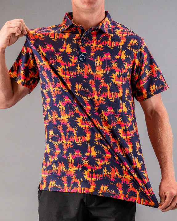 Men’s Palm Drip Dusk golf polo by Pins & Aces featuring navy palm tree silhouettes over a vibrant orange, pink, and purple tropical sunset print with an athletic fit.