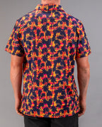 Men’s Palm Drip Dusk golf polo by Pins & Aces featuring navy palm tree silhouettes over a vibrant orange, pink, and purple tropical sunset print with an athletic fit.