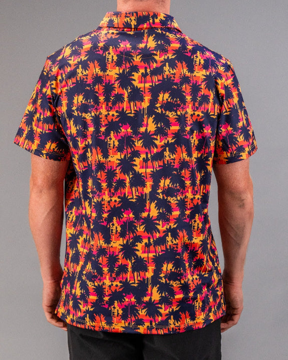 Men’s Palm Drip Dusk golf polo by Pins & Aces featuring navy palm tree silhouettes over a vibrant orange, pink, and purple tropical sunset print with an athletic fit.