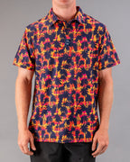 Men’s Palm Drip Dusk golf polo by Pins & Aces featuring navy palm tree silhouettes over a vibrant orange, pink, and purple tropical sunset print with an athletic fit.