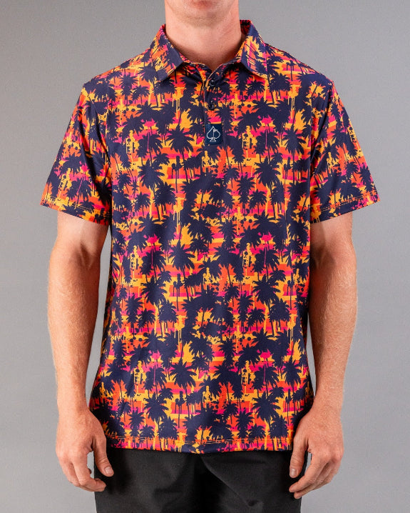 Men’s Palm Drip Dusk golf polo by Pins & Aces featuring navy palm tree silhouettes over a vibrant orange, pink, and purple tropical sunset print with an athletic fit.