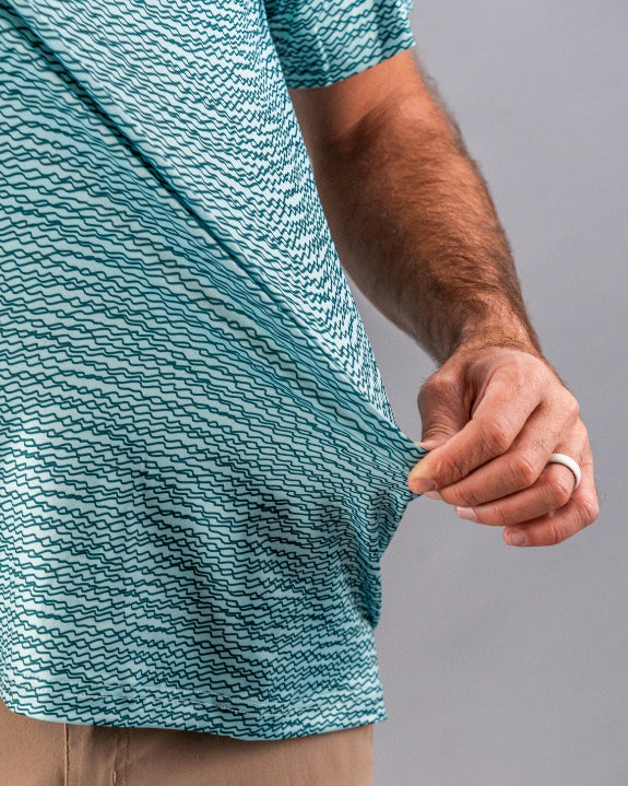 Men’s Squiggle Teal golf polo by Pins & Aces featuring an all-over aqua and teal abstract wavy squiggle pattern with short sleeves and an athletic performance fit.