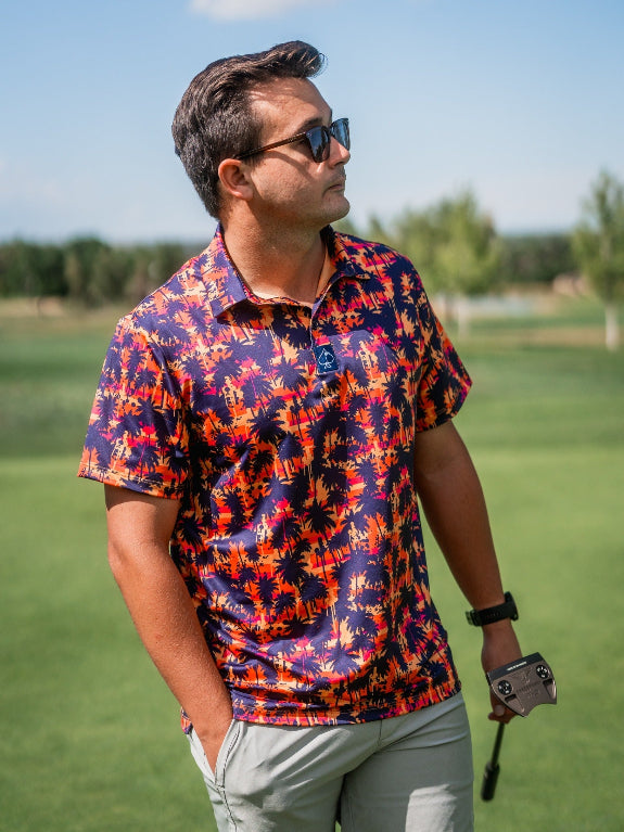 Men’s Palm Drip Dusk golf polo by Pins & Aces featuring navy palm tree silhouettes over a vibrant orange, pink, and purple tropical sunset print with an athletic fit.