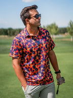 Men’s Palm Drip Dusk golf polo by Pins & Aces featuring navy palm tree silhouettes over a vibrant orange, pink, and purple tropical sunset print with an athletic fit.