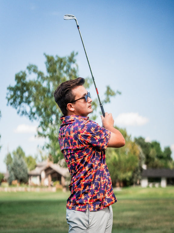 Men’s Palm Drip Dusk golf polo by Pins & Aces featuring navy palm tree silhouettes over a vibrant orange, pink, and purple tropical sunset print with an athletic fit.