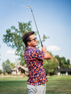 Men’s Palm Drip Dusk golf polo by Pins & Aces featuring navy palm tree silhouettes over a vibrant orange, pink, and purple tropical sunset print with an athletic fit.