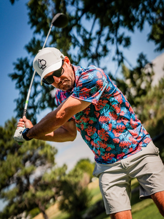 Men’s Painted Monstera Blue golf polo by Pins & Aces featuring coral and aqua tropical monstera leaves on a deep blue background with short sleeves and an athletic performance fit.