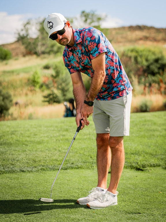 Men’s Painted Monstera Blue golf polo by Pins & Aces featuring coral and aqua tropical monstera leaves on a deep blue background with short sleeves and an athletic performance fit.