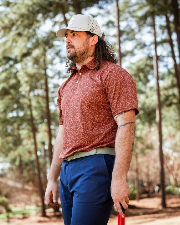 Men’s Filament golf polo by Pins & Aces featuring a rust and burnt orange abstract line pattern, short sleeves, modern blade collar, and athletic performance fit.