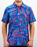 Men’s Cabo Colada golf polo by Pins & Aces featuring a vibrant coral red and aqua tropical floral print on a rich blue background with short sleeves and an athletic performance fit.