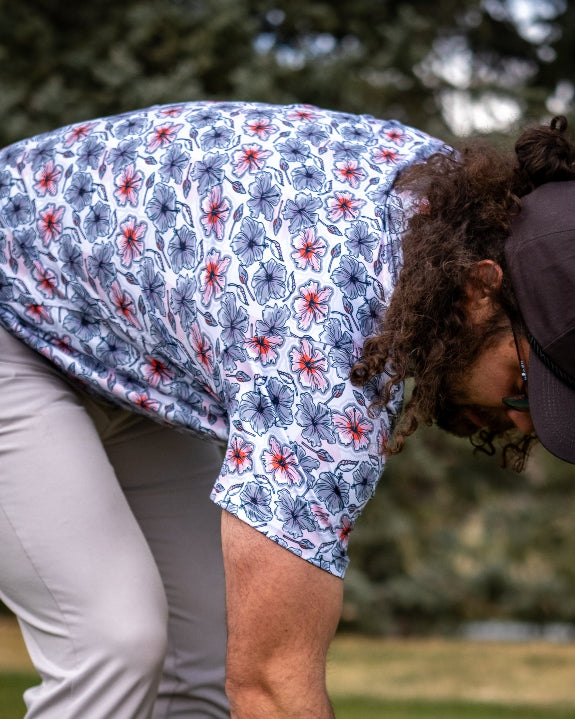 Men’s Hibiscus Harmony golf polo by Pins & Aces featuring blue and coral hibiscus flowers on a white lightweight performance golf shirt.