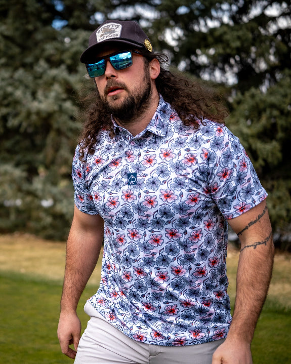 Men’s Hibiscus Harmony golf polo by Pins & Aces featuring blue and coral hibiscus flowers on a white lightweight performance golf shirt.