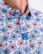 Men’s Hibiscus Harmony golf polo by Pins & Aces featuring blue and coral hibiscus flowers on a white lightweight performance golf shirt.