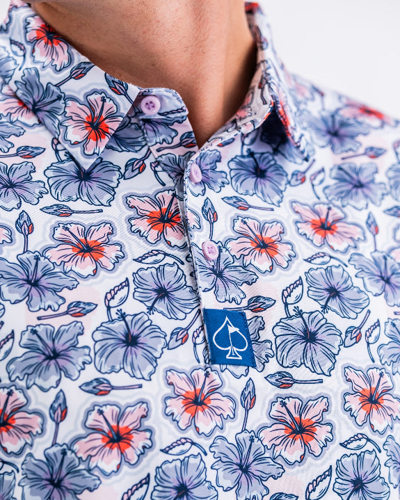 Men’s Hibiscus Harmony golf polo by Pins & Aces featuring blue and coral hibiscus flowers on a white lightweight performance golf shirt.