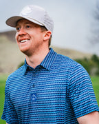 Men’s Rising Tides golf polo by Pins & Aces featuring a repeating blue and navy wave-inspired pattern, short sleeves, and an athletic performance fit.