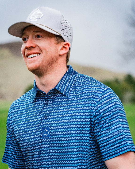 Men’s Rising Tides golf polo by Pins & Aces featuring a repeating blue and navy wave-inspired pattern, short sleeves, and an athletic performance fit.