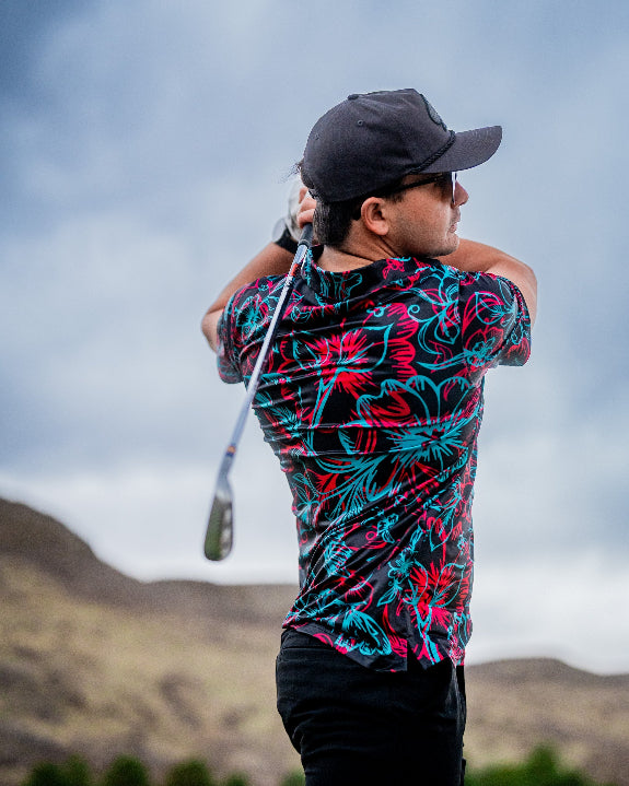 Men’s Cabo Electric golf polo by Pins & Aces featuring an aqua, hot pink, and coral tropical floral and leaf print on a dark charcoal background with an athletic performance fit.