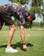Men’s Moon Burn golf polo by Pins & Aces featuring an abstract purple, blue, and black cosmic-inspired pattern with short sleeves and an athletic performance fit.