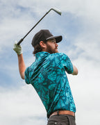 Men’s Cabana Nights golf polo by Pins & Aces featuring a blue and teal tropical leaf print, short sleeves, and athletic performance fit.
