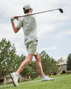Men’s Altitude Adjustment golf polo by Pins & Aces featuring an off-white base with a subtle blue-green mountain range print, short sleeves, and athletic performance fit.