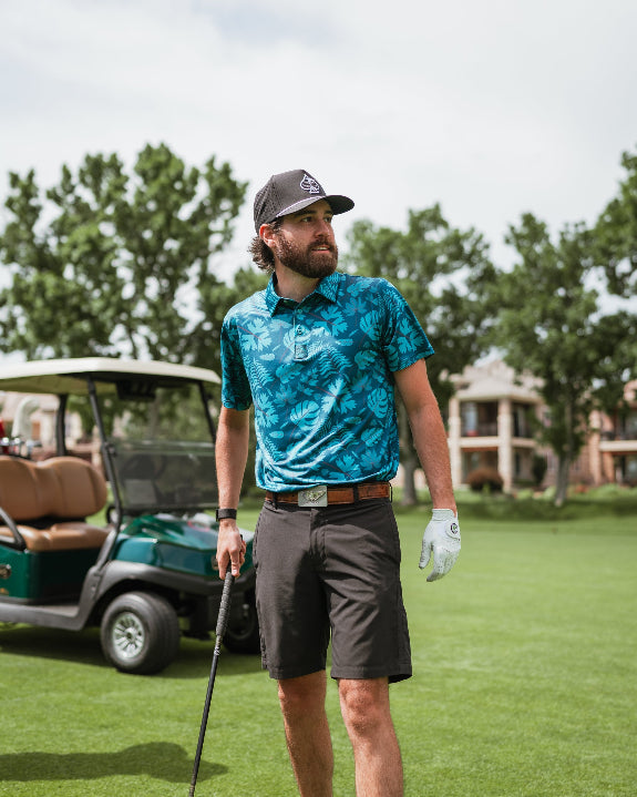 Men’s Cabana Nights golf polo by Pins & Aces featuring a blue and teal tropical leaf print, short sleeves, and athletic performance fit.