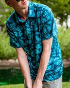 Men’s Cabana Nights golf polo by Pins & Aces featuring a blue and teal tropical leaf print, short sleeves, and athletic performance fit.