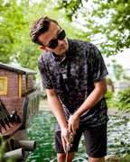Men’s Shadow Ops black camo golf polo by Pins & Aces featuring a tonal black and charcoal camouflage pattern, short sleeves, collared neckline, and athletic fit.