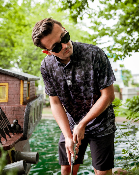 Men’s Shadow Ops black camo golf polo by Pins & Aces featuring a tonal black and charcoal camouflage pattern, short sleeves, collared neckline, and athletic fit.
