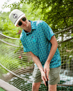 Men’s Fish & Drips golf polo by Pins & Aces featuring a bright aqua blue all-over fish print with colorful fishing-inspired details, short sleeves, and an athletic performance fit.