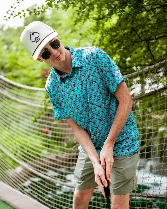 Men’s Fish & Drips golf polo by Pins & Aces featuring a bright aqua blue all-over fish print with colorful fishing-inspired details, short sleeves, and an athletic performance fit.