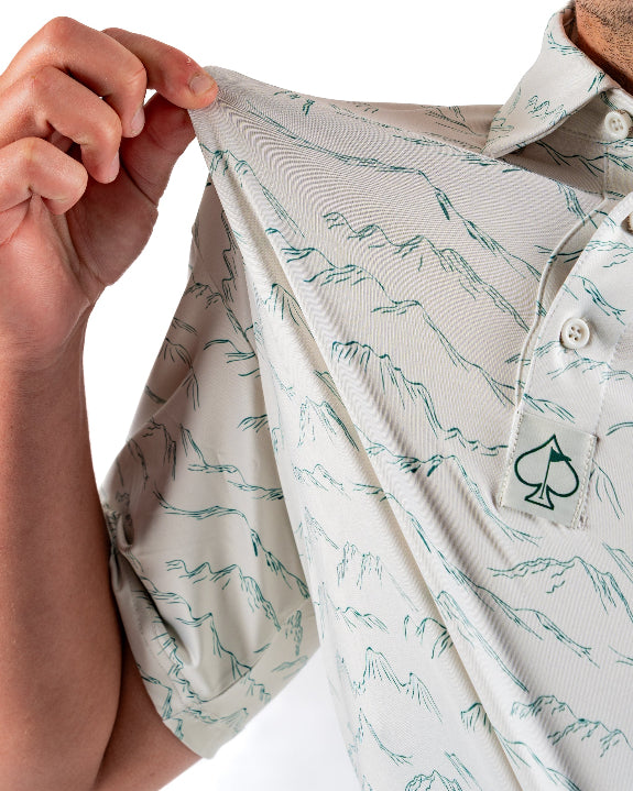 Men’s Altitude Adjustment golf polo by Pins & Aces featuring an off-white base with a subtle blue-green mountain range print, short sleeves, and athletic performance fit.