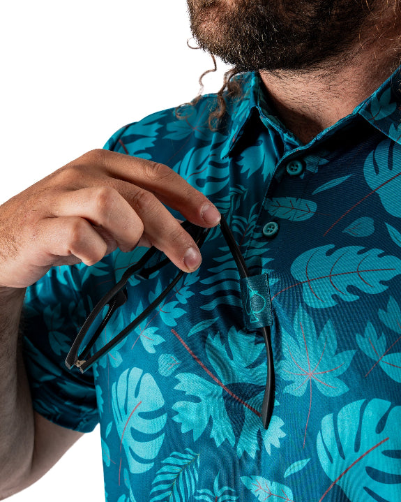 Men’s Cabana Nights golf polo by Pins & Aces featuring a blue and teal tropical leaf print, short sleeves, and athletic performance fit.