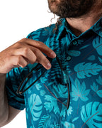 Men’s Cabana Nights golf polo by Pins & Aces featuring a blue and teal tropical leaf print, short sleeves, and athletic performance fit.